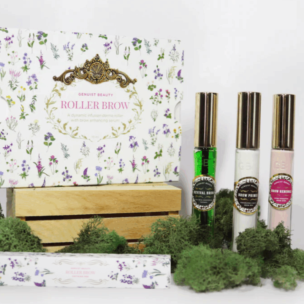 Display of Genuist Beauty’s Total Brow Care System, including Roller Brow derma roller with serum, Revival Brow daily conditioner, Brow Primer, and Brow Renewal nightly treatment. Products are styled with floral packaging, greenery, and a wooden crate accent.